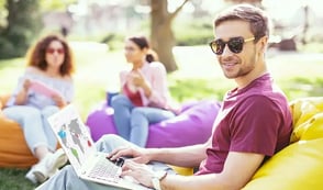 A man is working outside, sitting on a yellow bean bag and has his laptop on his lap. He has sunglasses on and is enjoying working in the sunshine. Behind him are his two colleagues who are also sat on bean bags and working.